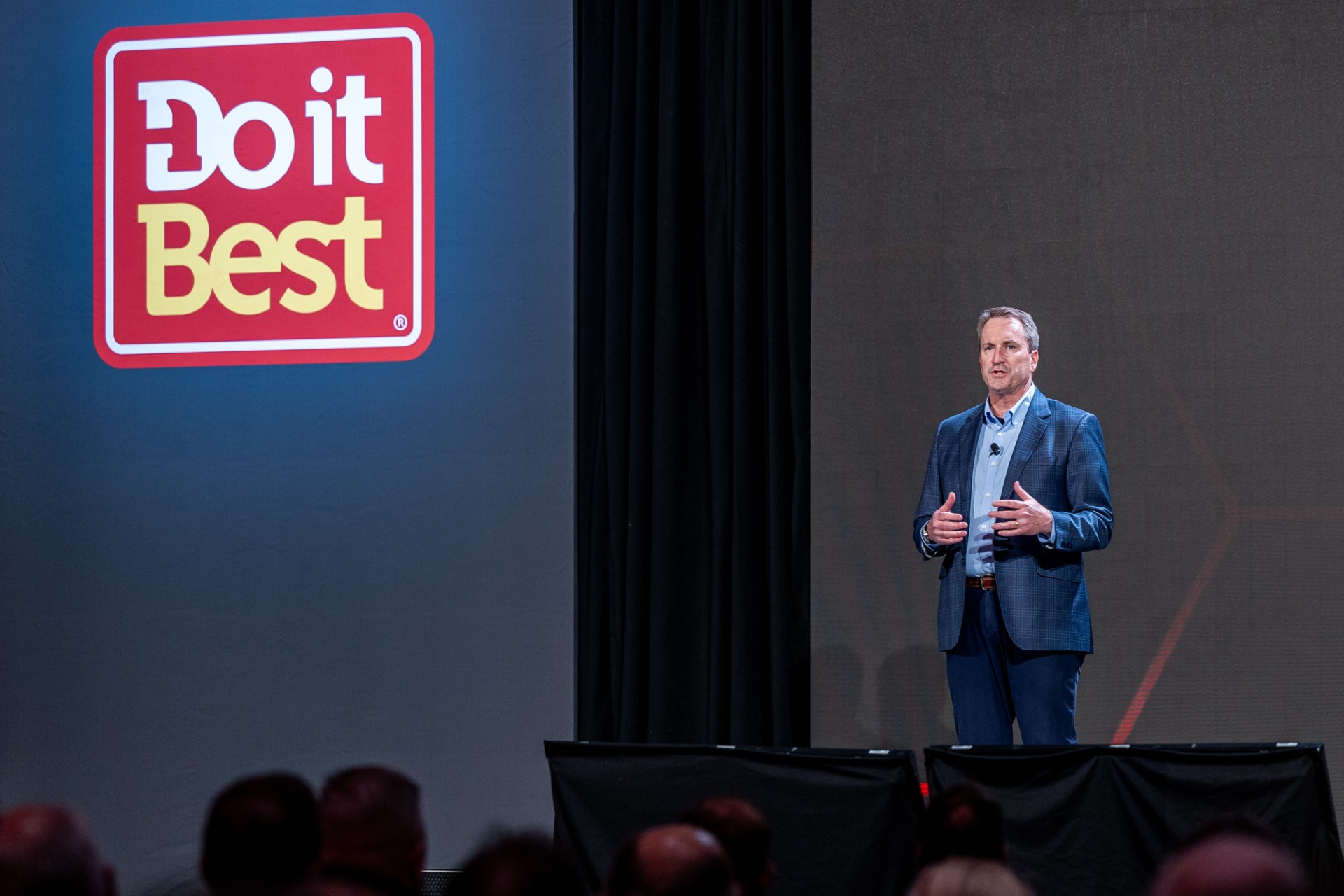 Male speaker in a blue checkered suit presenting on stage beside a large Doit Best logo on the left wall.
